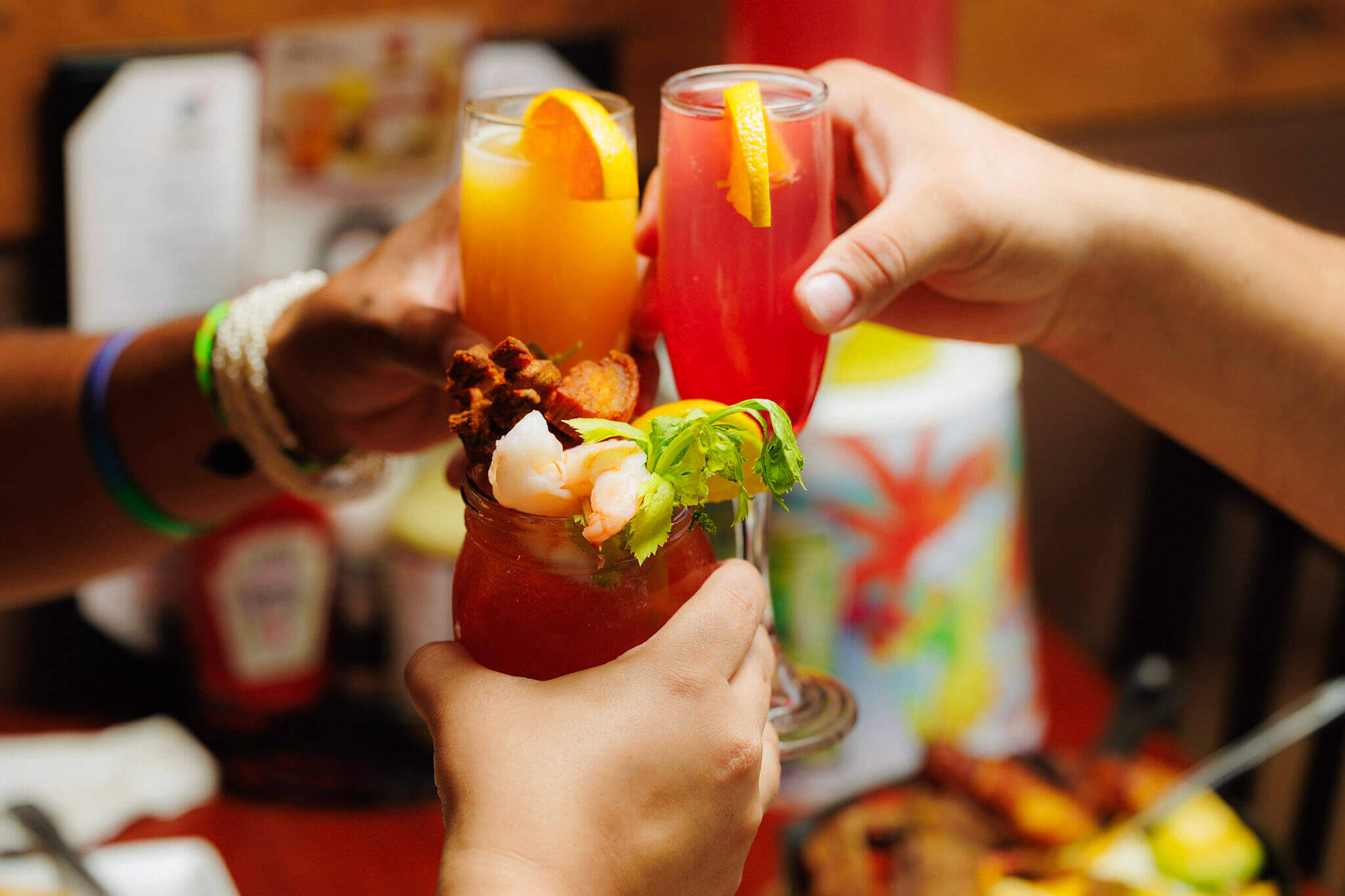 Three people toasting with a bloody mary, an orange juice, and a pink cocktail.