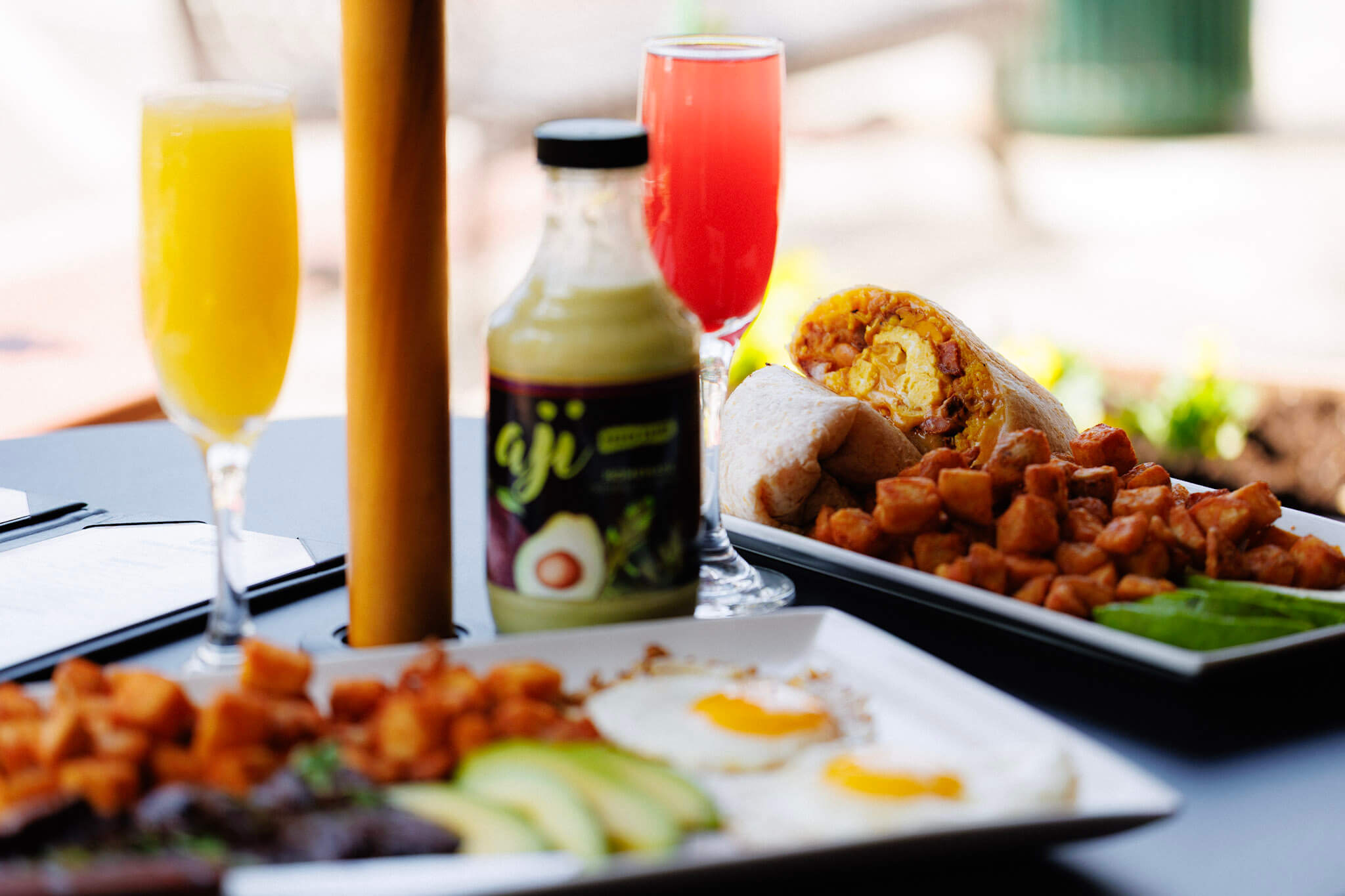 Breakfast plates featuring breakfast burritos, fried eggs, avocado slices, and potatoes, accompanied by two glasses of juice and a bottle of Aji sauce.