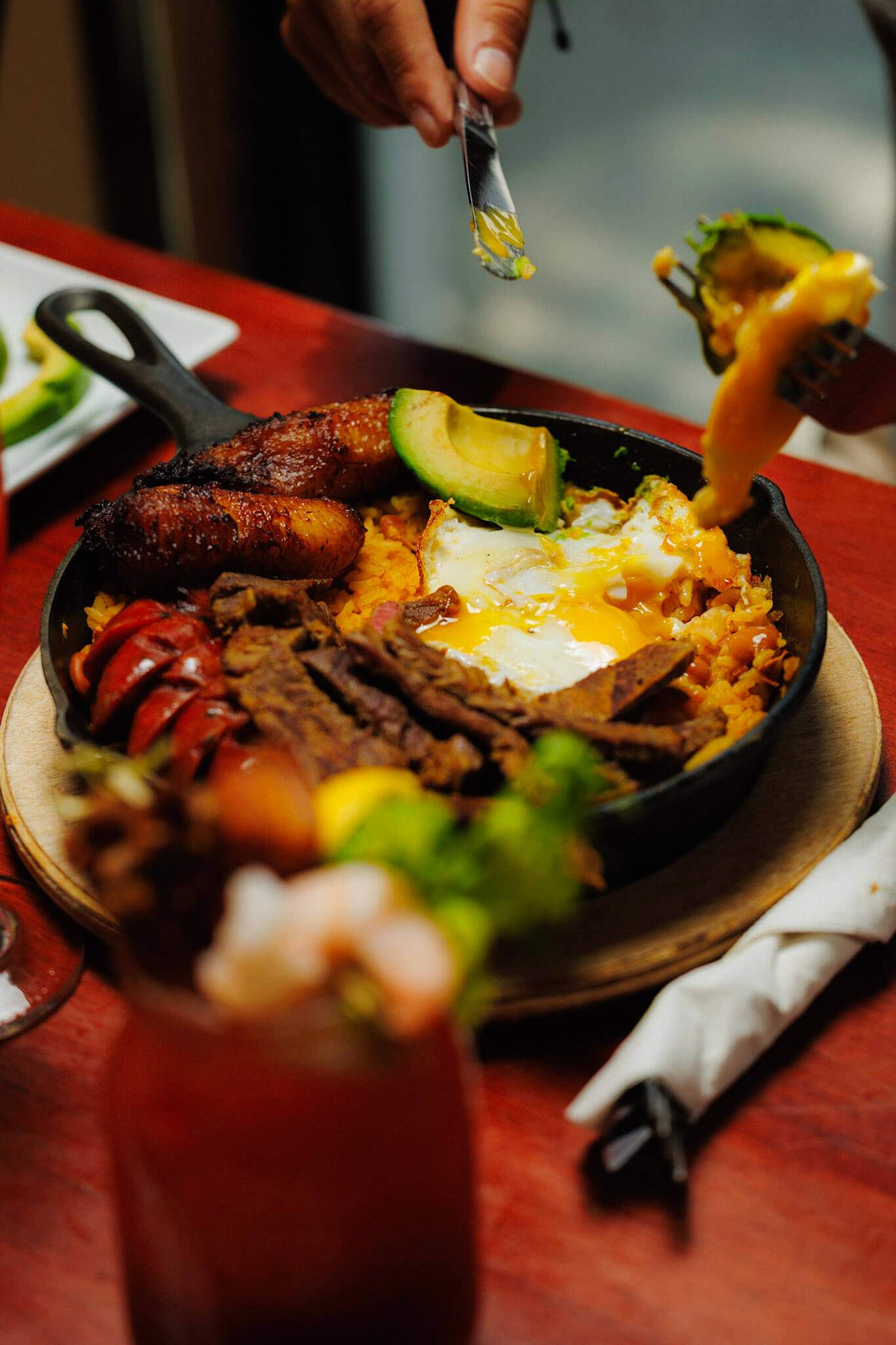 A skillet meal of rice, fried egg, sliced meat, sausage, and avocado, with a hand using a fork to lift a piece of egg.