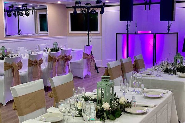 A banquet hall set for an event with decorated tables, white chairs with burlap bows, glassware, green lantern centerpieces, and purple uplighting near a DJ booth.