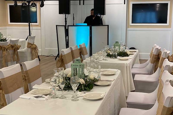 Two banquet tables with white tablecloths are set for a formal event, decorated with floral centerpieces and tan chair sashes. A DJ booth with blue lighting is in the background.