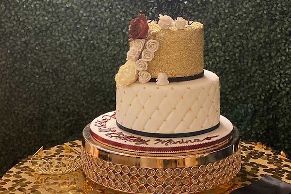 A three-tiered birthday cake with gold glitter, white quilted design, white and red roses, and a decorative base, displayed on a gold-patterned table.