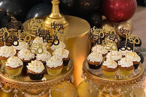 Cupcakes with white frosting and gold sprinkles, each topped with a “cheers to 50 years” sign, arranged on decorated gold stands with black and gold balloons in the background.