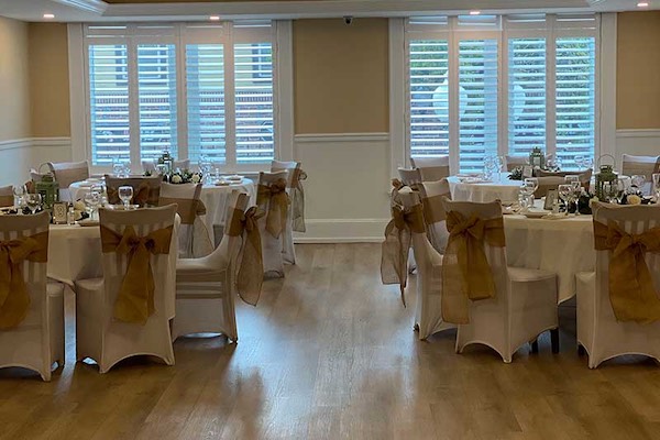 A banquet room with round tables set for an event, chairs covered in white fabric with brown bows, and large windows with white shutters in the background.