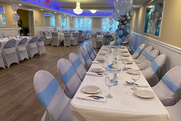 A banquet hall is set up for an event with tables covered in white cloths, blue and white balloons, and white chairs with blue sashes. The room is well-lit and decorated.