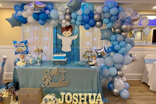 A party table decorated with blue, white, and silver balloons, a cake, angel cutout, treats, and a large name sign reading "Joshua" for a baptism celebration.
