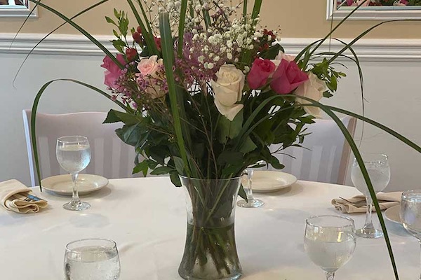 A glass vase with pink, white, and purple flowers and greenery sits on a round table set with glasses of water, plates, and folded napkins.