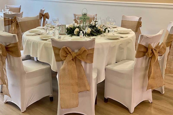 Round table set for a formal event with white tablecloth, white chair covers, and burlap bows; centerpiece with greenery and flowers.