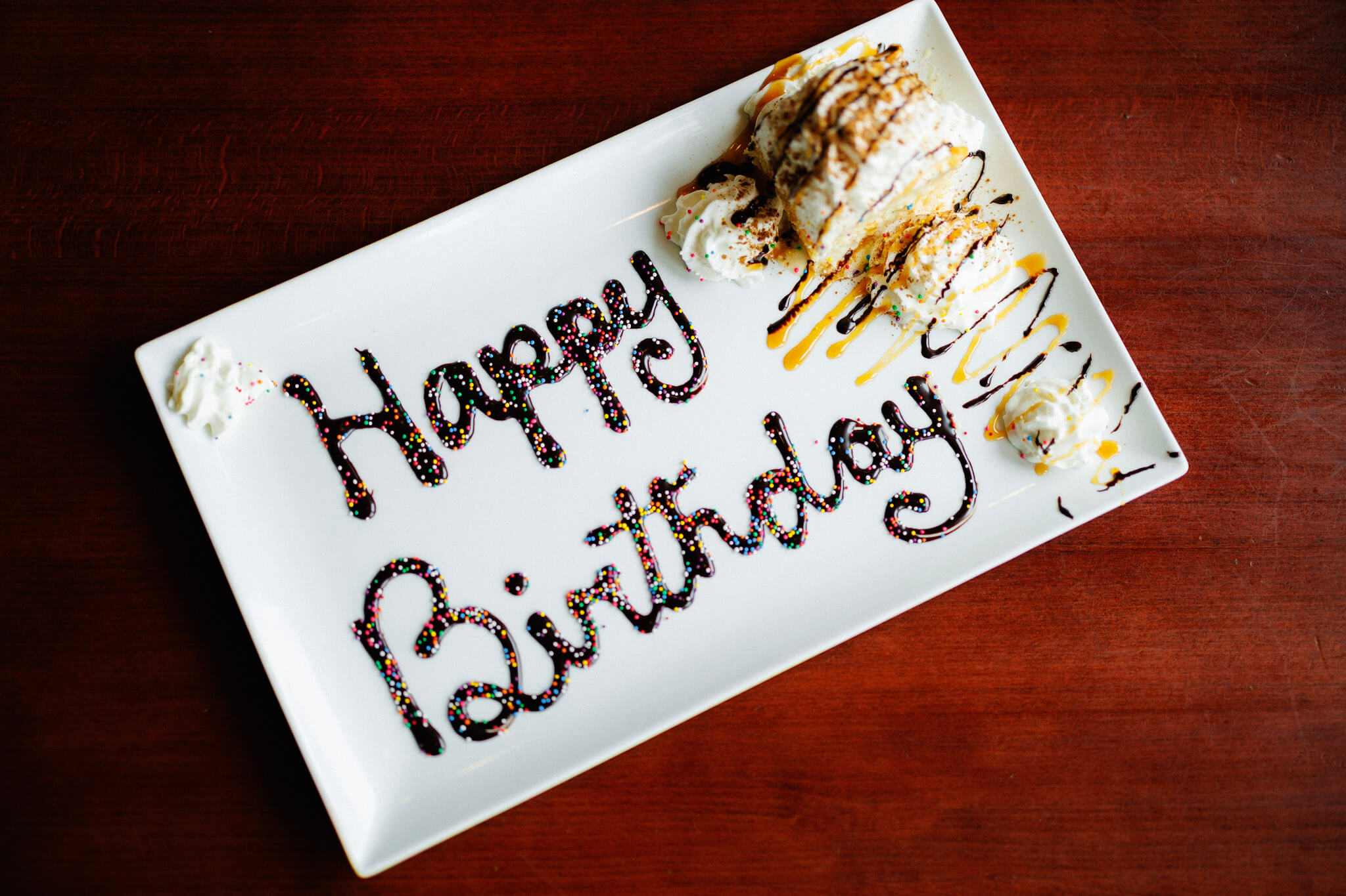 Rectangular plate with “Happy Birthday” written in chocolate syrup and sprinkles, served with a pastry and whipped cream.