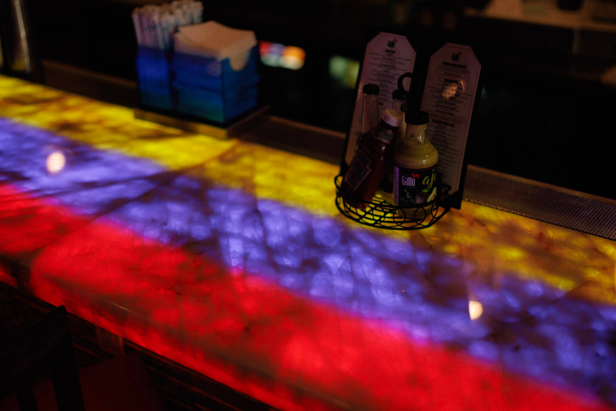 A bar counter illuminated with red, yellow, and blue light, featuring a caddy holding condiment bottles and menus.