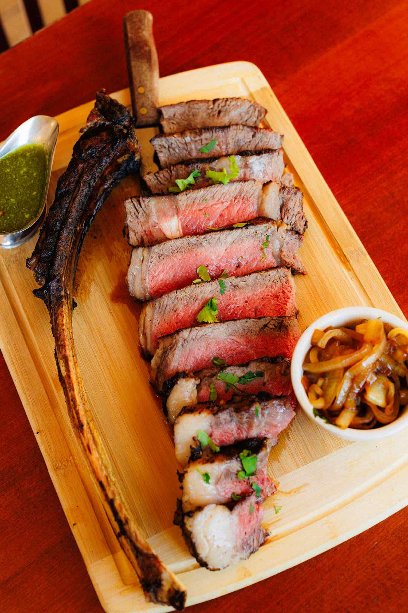 Sliced tomahawk steak served on a wooden board with a small bowl of sautéed onions and a side of green sauce.