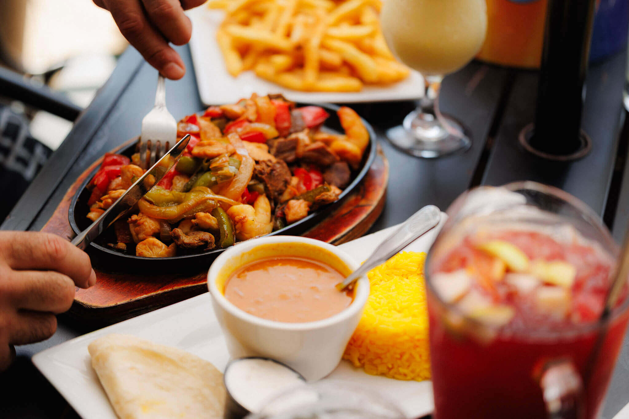 A person eating a sizzling skillet of fajitas, served with a side of rice, beans, a tortilla, and drinks.