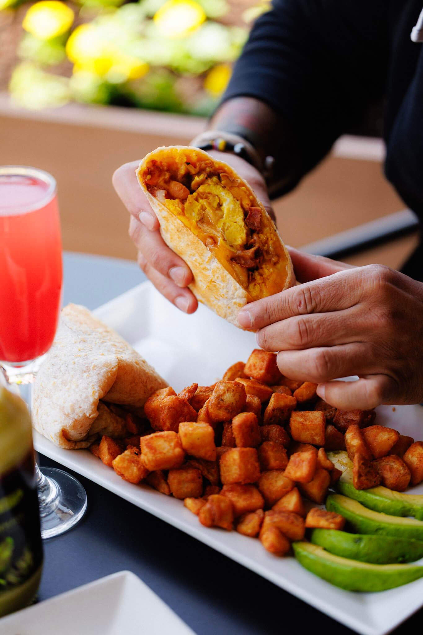 A person holding a breakfast burrito filled with eggs, cheese, and meat, with a side of crispy potatoes and avocado slices on a white plate.