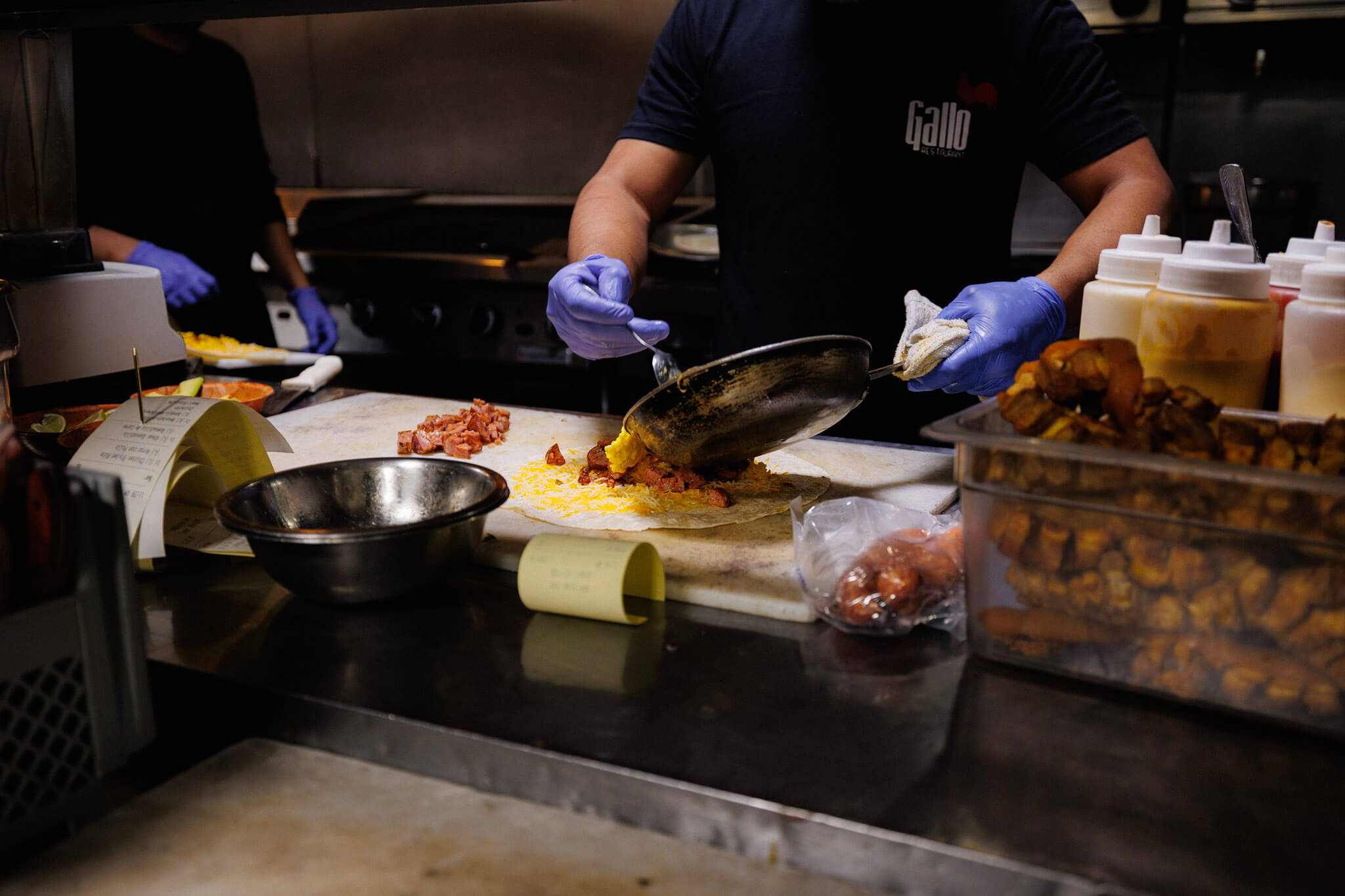 A cook in blue gloves using a spatula to empty a pan of cooked eggs and meat onto a flour tortilla on a restaurant prep counter.