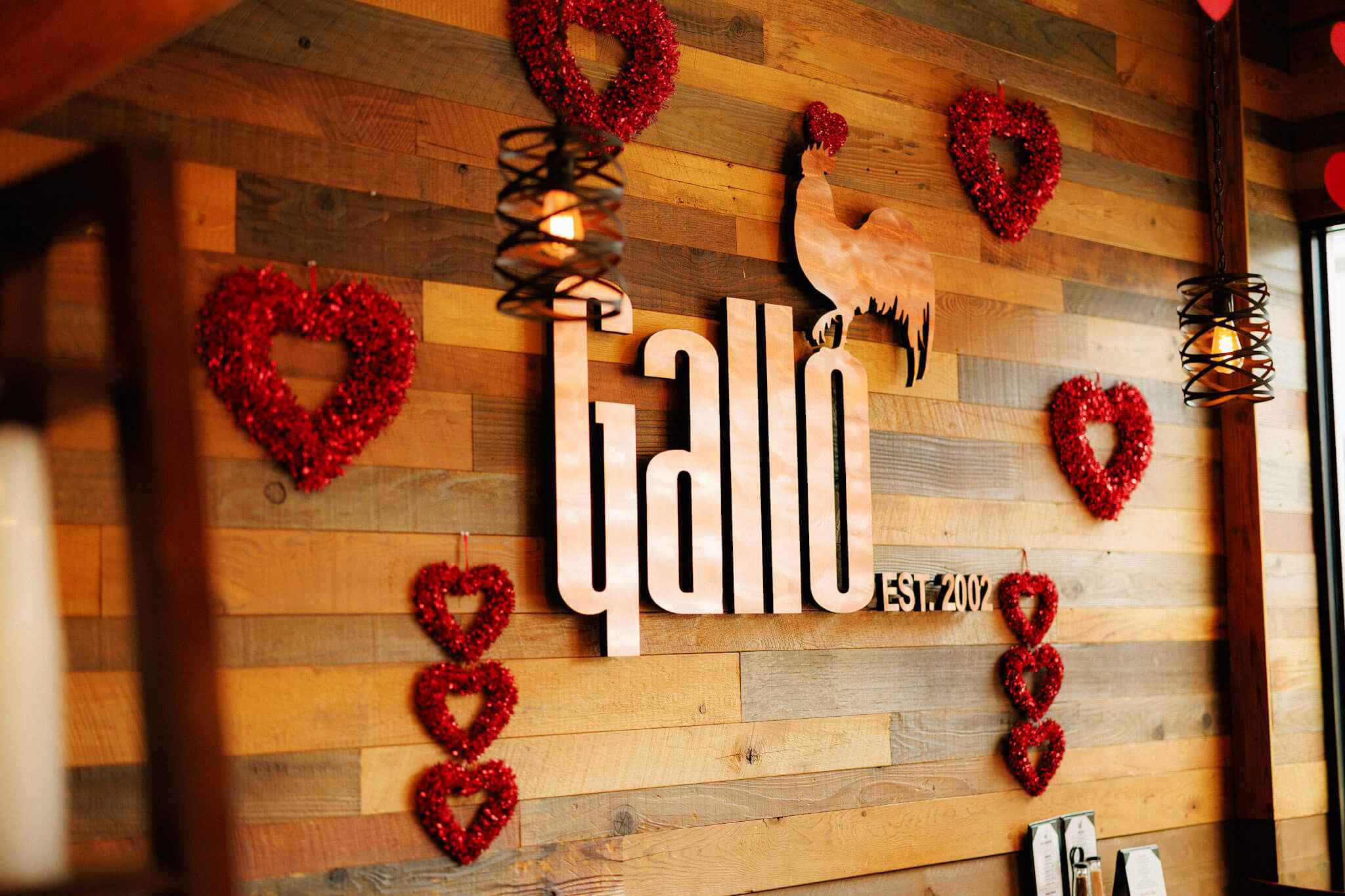 The “Gallo” restaurant logo and “Est. 2002” text mounted on a wooden wall decorated with red heart-shaped wreaths.