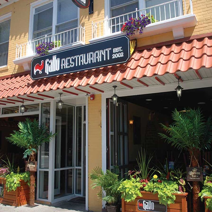Exterior view of Gallo Restaurant with red tile roof, potted plants, and outdoor seating under a covered patio. Signs display the restaurant's name and “Est. 2002.”.
