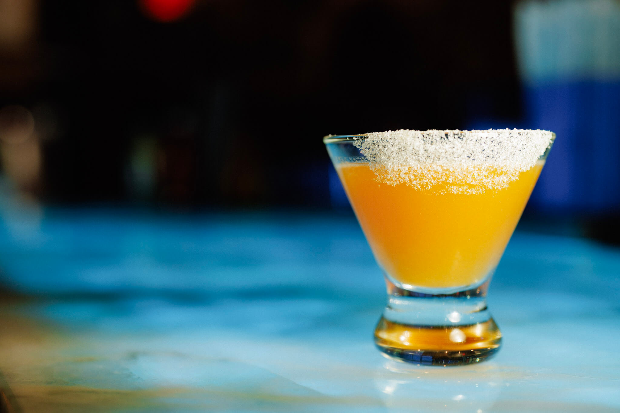 An orange cocktail in a glass with a sugar-rimmed edge, sitting on a blue surface.