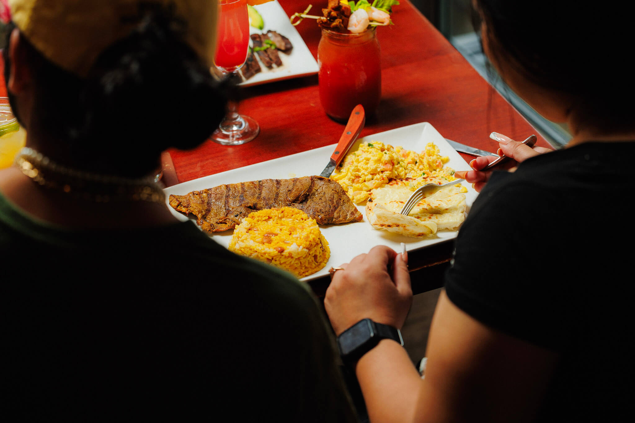 Two people eating a meal of grilled steak, yellow rice, scrambled eggs, and arepas at a restaurant table.