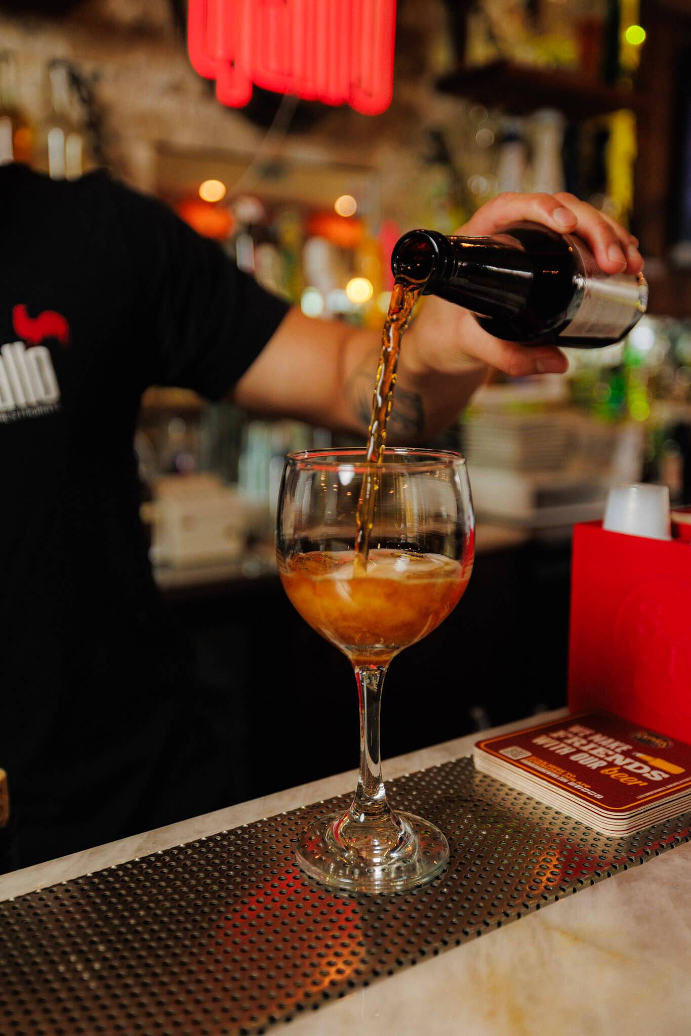 A bartender pours beer from a glass bottle into a stemmed glass on a bar counter.