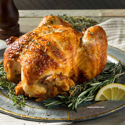 A roasted chicken served on a metal plate, garnished with fresh rosemary, thyme, and a lemon wedge.