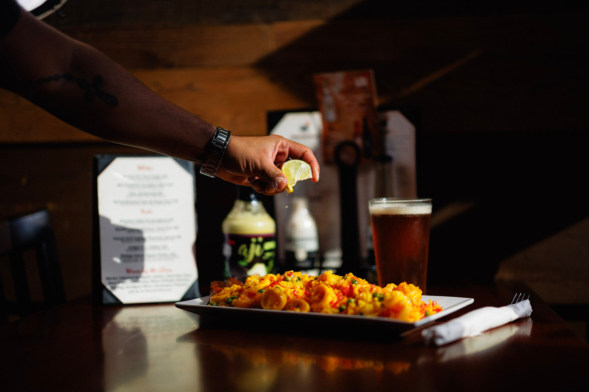 A hand squeezes a lime wedge over a plate of yellow seafood paella, with a glass of beer and a menu on the table.