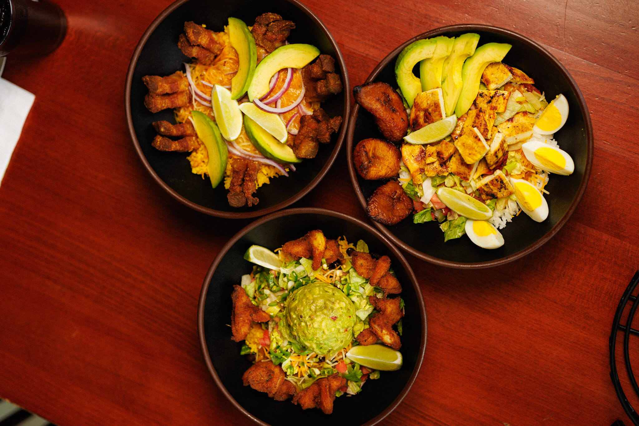 Three bowls of food served on a wooden table: one with pork and avocado, one with chicken, hard-boiled eggs, and plantains, and one with shrimp and a scoop of guacamole.