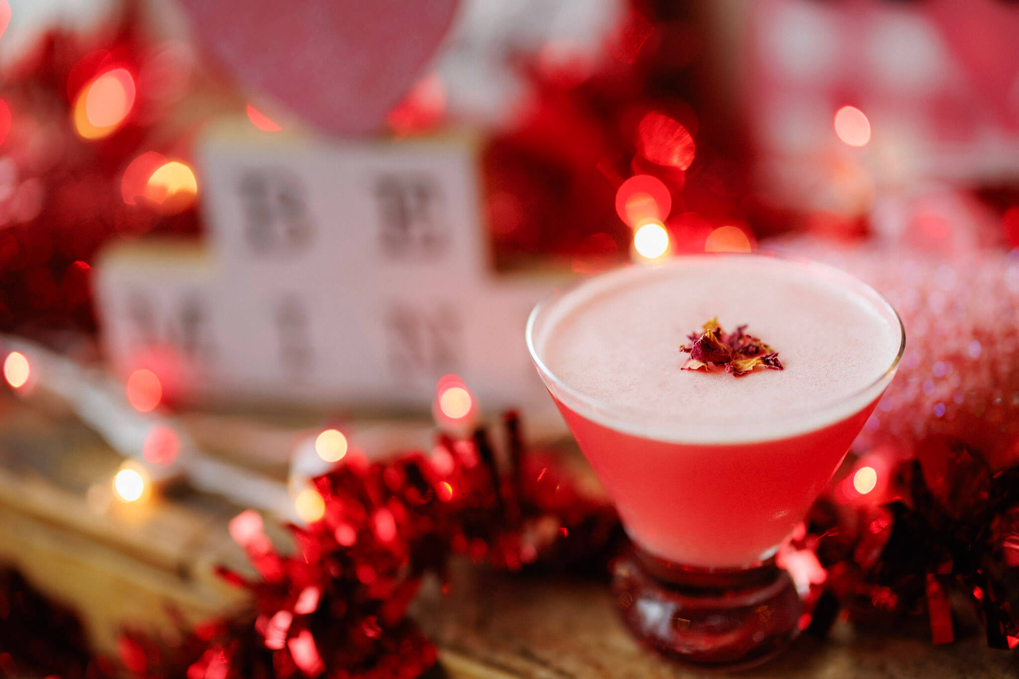 A pink cocktail in a coupe glass topped with dried flower petals, surrounded by red tinsel and blurred Valentine’s Day decorations.
